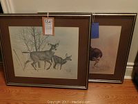 Two mounted Glen Loates wildlife prints leaning against a wall, with labels attached. One print features two deer, the other a moose.