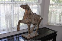 Full view of carved wood horse statue mounted on pedestal placed on a black furniture piece against a window with lace curtains.