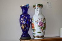 Photo of two vases side by side, one white porcelain with peacock motif, and one blue glass floral vase on wooden stand.