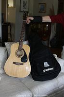 Full view of Yamaha SCF08 acoustic guitar standing upright next to its black Yamaha-branded carrying case on a couch.