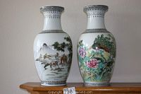 Two Asian porcelain vases side by side on wooden surface against beige wall. One vase illustrated with colorful peacock among flowers, the other depicting horses grazing under a tree.