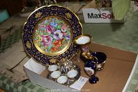 Photo showing a collection of porcelain including a large decorative plate with floral design, small cups, and vases with gold accents, placed in a cardboard box.