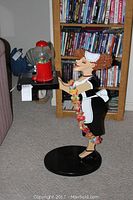Full view of Millie the Maid figurine holding a tray with red gumball machine, positioned on carpet in front of bookshelf.