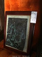 Framed metal relief artwork of sailing ships leaning against a chair.