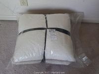 Two sheepskin wool pillows tightly packed together in protective clear plastic packaging with black straps at midsection.