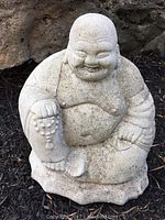 Front angled view of a garden Buddha statue on soil next to rocks, showing wear and soil marks on the stone surface.