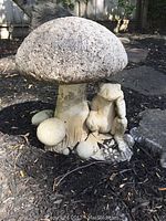Concrete toadstool mushroom yard ornament with smaller mushrooms and crouching frog attached, shown in garden bed with mulch.