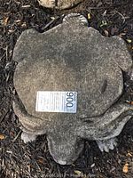 Close-up of a weathered concrete stepping stone shaped like a rabbit, showing natural patina and soil on the surface, with a lot tag.