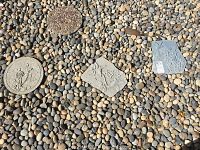 Three decorative stepping stones laying on small garden rocks. One round stone with floral design, two irregular shaped stones with bird motifs.