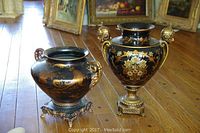 Pair of black urns with gold floral decoration and ornate detailing on wooden floor with paintings in background. Larger urn has figure handles, smaller urn has curled handles and decorative stand.