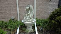 Front view of white cherub fountain against brick wall with landscape foliage