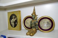 Buddha statue in kneeling position flanked by two oval framed Asian art prints and one rectangular framed print of woman with horse.