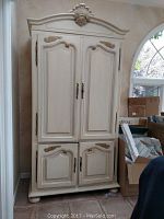 Closed view of cream wooden armoire with gold accents, four doors with decorative molding and metal handles, placed in corner of room.