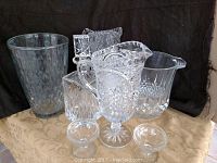 Photo showing two pitchers, ice bucket, vase, crystal decanter, and two small pedestal glass bowls arranged on a cloth surface.