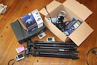 Wide shot of the lot showing two black Sunpak tripods, Sony Handycam box, Zenith VCR, two digital cameras (Sony Cybershot and Kodak EasyShare), and various cables and accessories in cardboard boxes on wooden floor.