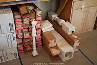Several white porch railing balusters and large columns stacked on a floor with many original cardboard boxes behind them.
