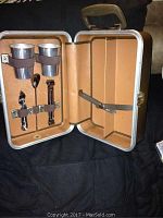 Open tan aluminum case showing two metal martini shaker cups held with brown elastic straps inside the left panel and empty right panel with a strap.