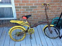 Side view of the folding bicycle showing brown frame, yellow rims, and rear rack, black and red saddle, flat tires