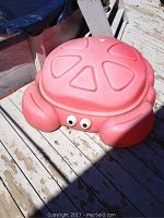 Red turtle shaped sandbox with faded red plastic and cartoon eyes on side, shown on a wooden deck.