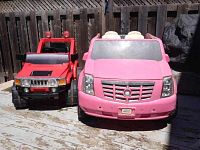 Both pink and red kids battery operated ride-on cars shown side by side outdoors on wooden deck.