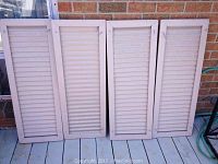 Four reclaimed aluminum shutters leaning against a brick wall on a deck, showing horizontal slats and white paint with signs of wear.