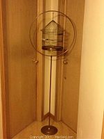 Full view of brass birdcage hanging inside the circular brass stand in a corner.