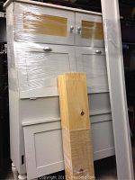 Front view of wrapped wooden bed frame including glass door display and storage compartments, with additional wooden boards leaning on it.