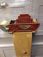 Side view of red cast iron device with brush attached, showing metal clip holding rolled sheets on left side and stepped design center.