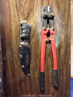 Bolt cutter and jin knife with case shown on wooden surface, bolt cutter has visible rust on cutting head, red handles with textured black grips, jin knife in black sheath with snap closure and brass accent on handle.