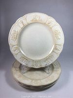 Four white antique Wedgwood dinner plates stacked with one displayed upright showing fluted rim and cherub motifs on border.