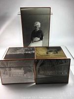 Five glass and copper photographic negatives displayed on a white surface; includes one large vertical glass negative of a seated woman and four horizontal architectural scenes of municipal buildings and homes in different seasons.