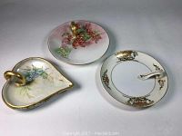 Three porcelain hand handled serving dishes: heart-shaped dish with blue floral design, round dish with grapes, and Noritake dish with gold pattern