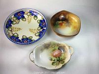 Three hand painted Nippon porcelain bowls shown from top, including blue floral, brownish footed bowl with fruit and flower motif, and white oval handled floral candy dish