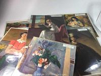 Photo showing multiple prints stacked, with visible artwork including woman reading by window, portrait of woman, and floral still life.