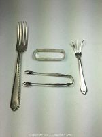 Four sterling silver pieces displayed on a flat surface: large dinner fork with ornate Lady Hilton pattern, sugar tongs, olive fork, and oval napkin ring.