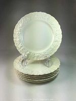 Six cream Wedgwood Queensware salad plates stacked with one elevated in a display stand showing off embossed design and fluted edges.