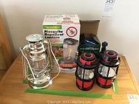 Lot items arranged on table including a ThermaCELL Mosquito Repellent Lantern box, silver metal lantern, two red and black mini lanterns, and a blue flashlight partially seen on the left.