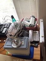 Multiple landline phones, plastic bins, and other small items on a wooden table near window blinds.