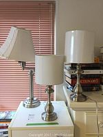 Photo showing three desk lamps on a table by a window with blinds. Two lamps have brushed metal bases and cylindrical white shades, the third has an adjustable arm with a rectangular shade.