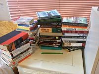 Photo showing multiple stacks of books placed on a surface near a window with blinds. Includes various titles and genres.