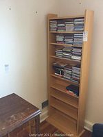 Full view of the wooden media shelf with CDs on 3 of the 5 shelves, empty bottom shelves visible, shelf stands in a corner beside a wooden table.