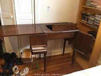 Wide angle view of the wooden sewing table with the fold-out extension open and a portion of the Singer machine compartment visible. The table is placed against a wall on a hardwood floor.