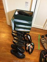 Multiple pairs of girls' black leather shoes including dress shoes and tap shoes arranged on wooden floor next to shoe boxes.