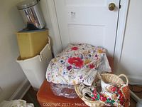 Orange ottoman with colorful embroidered linens on top, plastic bins stacked behind, and wicker basket nearby.