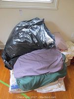 Black bag of pillows or quilts on top of a stack of folded bed linens in various colors on the floor near a window.