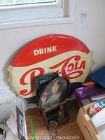 Photo of large Pepsi-Cola metal advertising sign with vintage red and cream colors, Coca Cola tin tray, and several vintage bottles grouped on the floor.