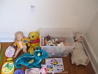 Three dolls, two plush toys, and a large plastic bin containing small furniture and dollhouse accessories set against a wall