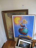 Photo showing a large framed landscape print and a colorful framed print of a child with globe and moon leaning against a wall.