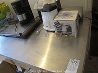 Wide view of kitchen appliances displayed on stainless steel surface including coffee maker, popcorn maker, fondue pot box, and Hot Tray warmer.