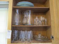 Overview of three large glass flower vases, assorted drinking glasses on two shelves inside a wooden cabinet